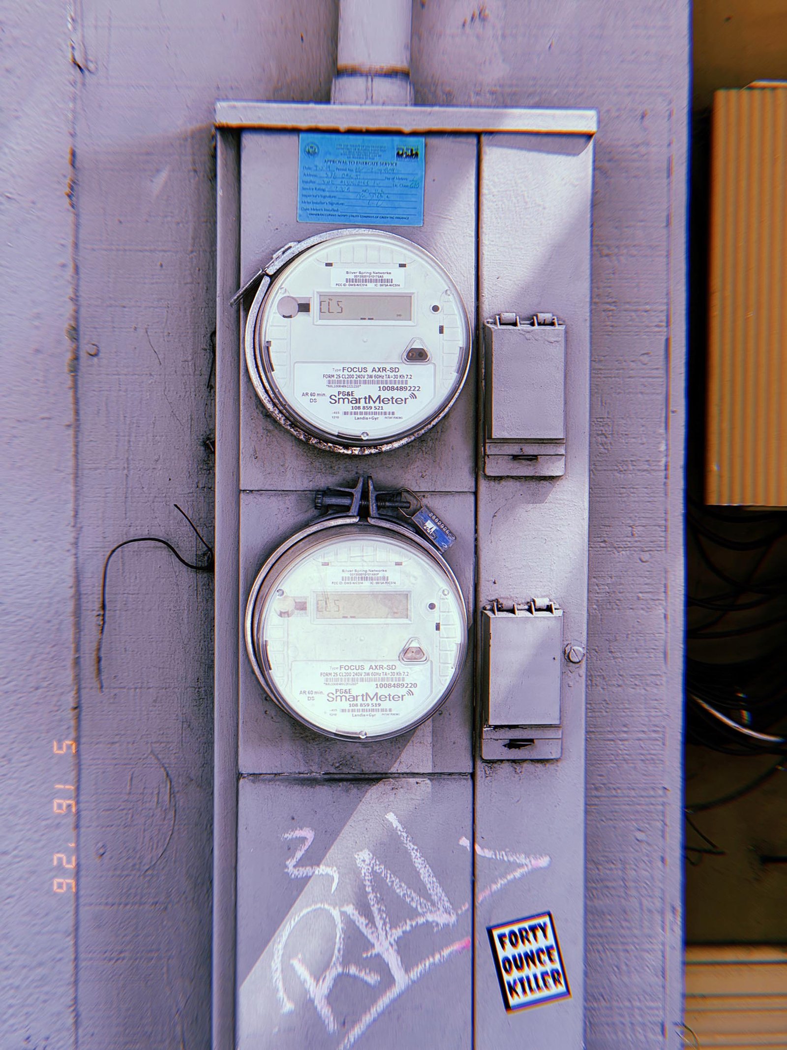 Two digital electrical smart meters mounted vertically within a utility service panel, with protective covers, electrical enclosures, graffiti markings, and a sticker attached to the lower section of the cabinet.