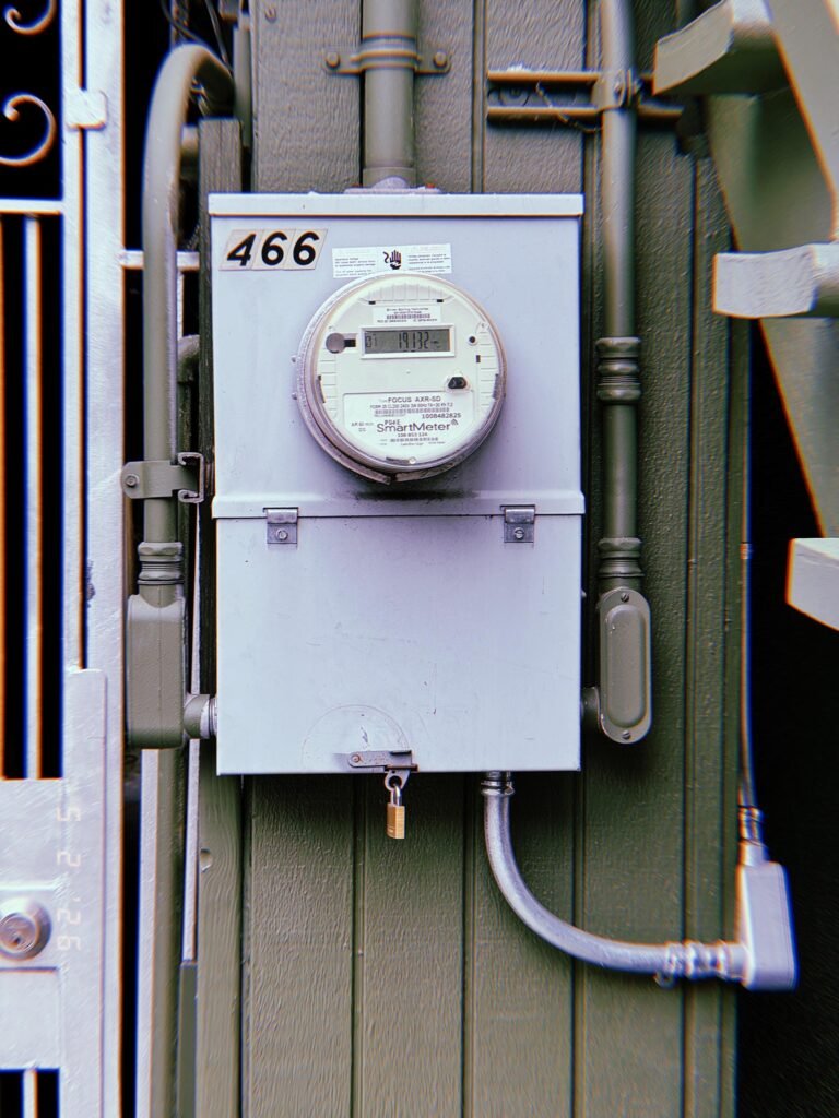 A digital electrical smart meter mounted on a locked utility service cabinet attached to a building exterior, connected through metal conduit and electrical distribution infrastructure.