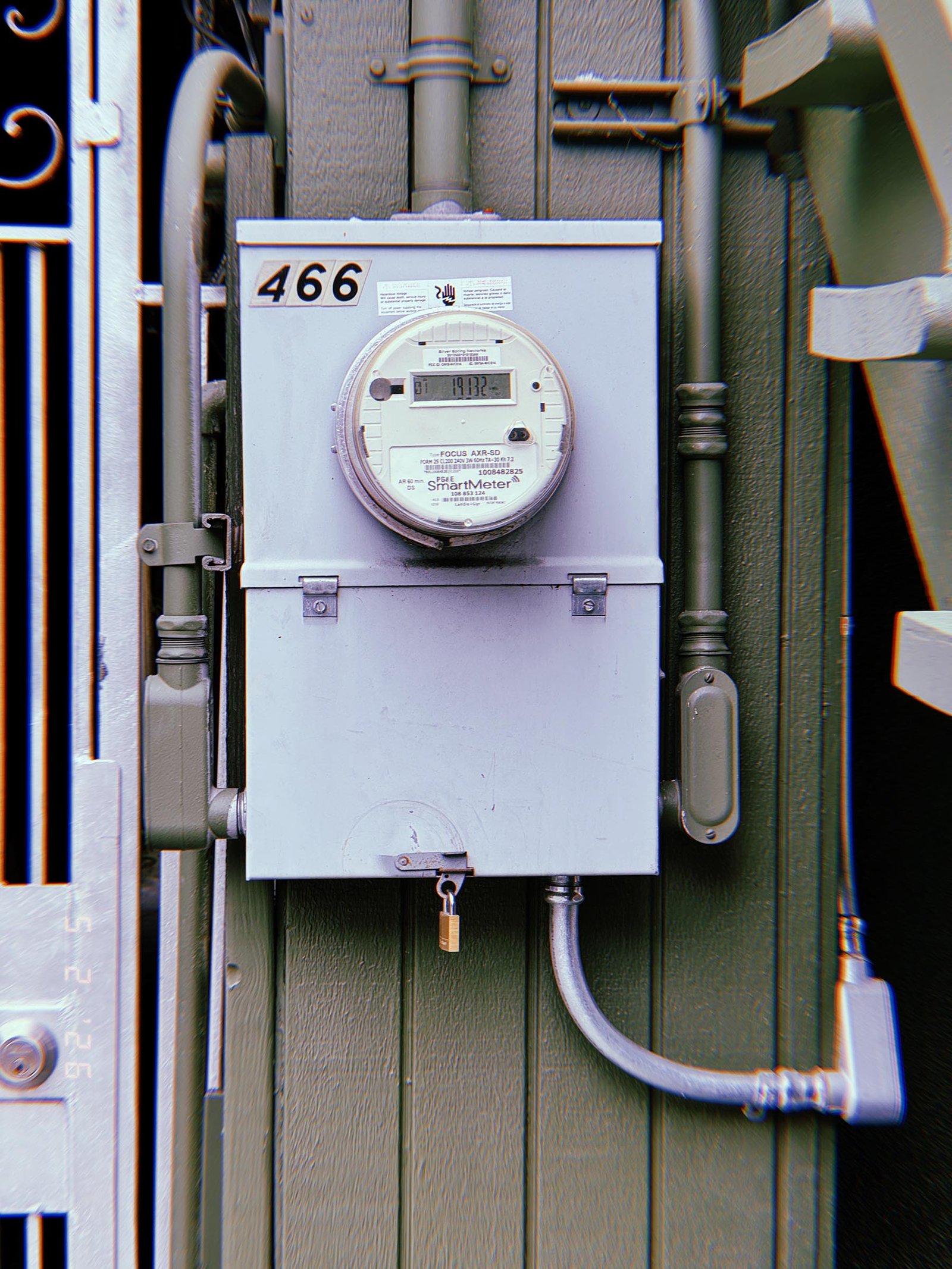 A digital electrical smart meter mounted on a locked utility service cabinet attached to a building exterior, connected through metal conduit and electrical distribution infrastructure.