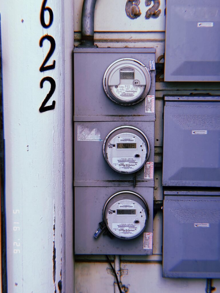 Three digital electrical smart meters mounted vertically within a multi-unit service panel, installed beside electrical distribution cabinets and building identification numbers.