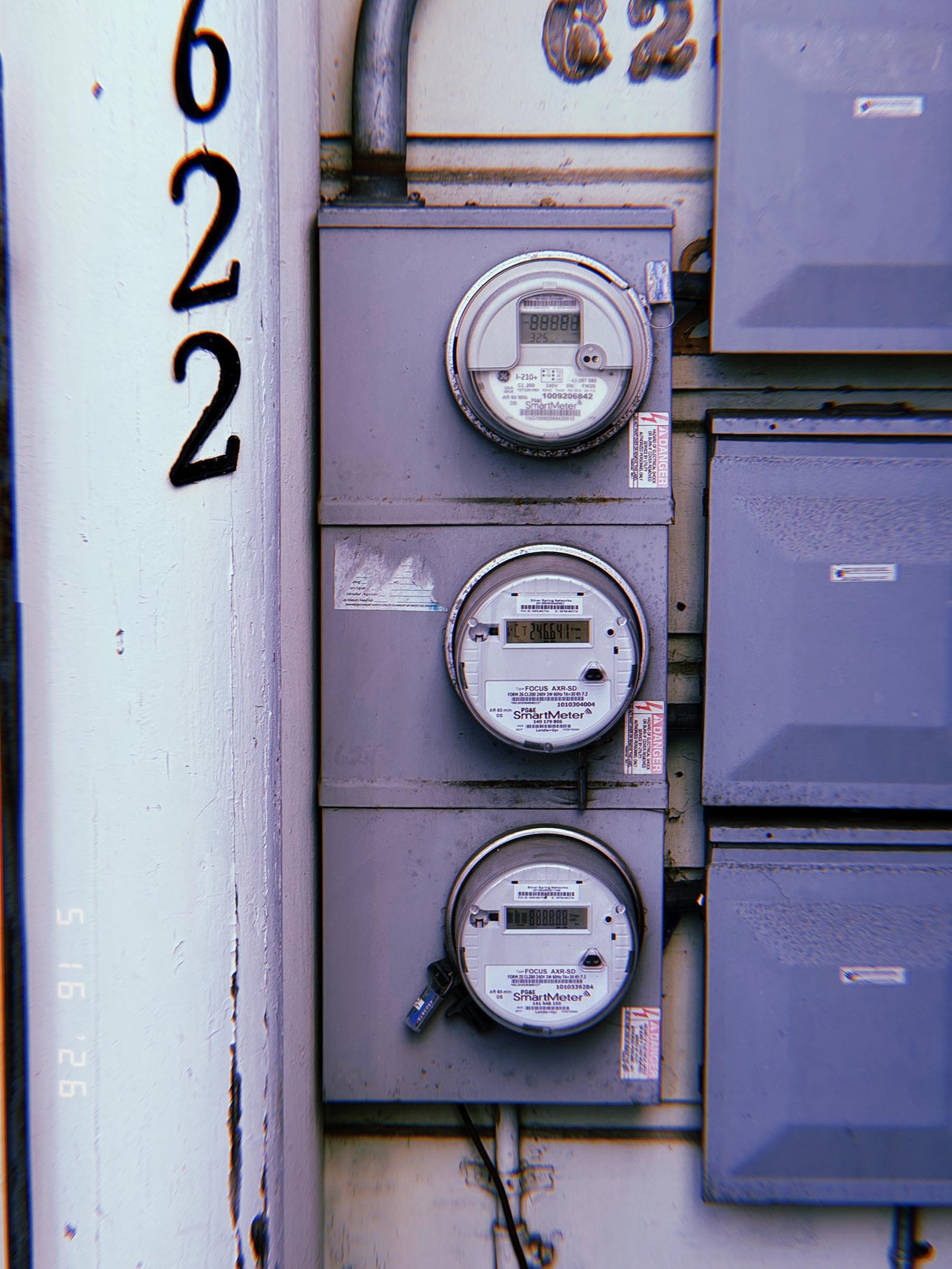 Three digital electrical smart meters mounted vertically within a multi-unit service panel, installed beside electrical distribution cabinets and building identification numbers.