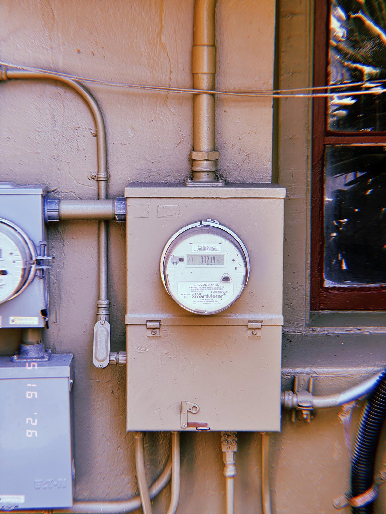A digital electrical smart meter mounted on a painted utility cabinet attached to a building wall, connected by metal conduit and service lines beside a window.