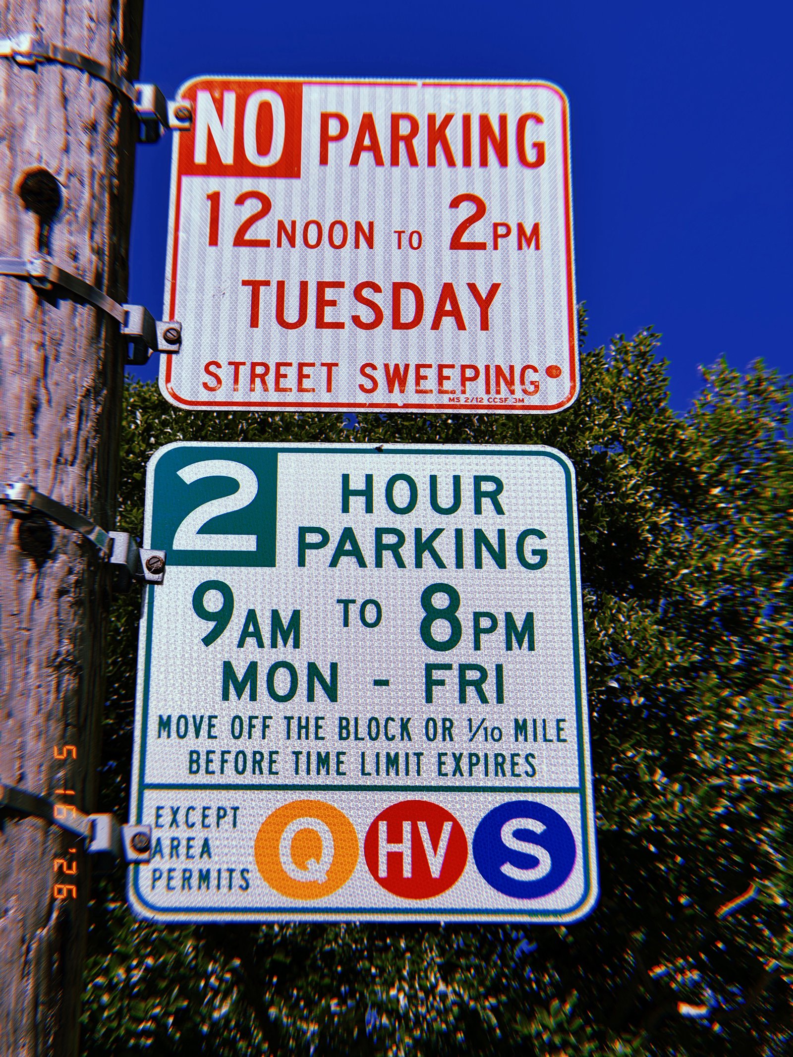 Street regulation signs mounted on a utility pole against a deep blue sky. The upper sign reserves curb access during scheduled street sweeping hours, while the lower sign imposes a two-hour parking limit and identifies permit-holder exemptions. Together they define the conditions governing vehicle occupancy within the curbside system.