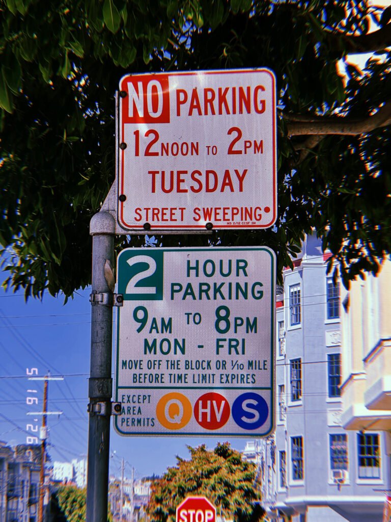 Municipal parking regulation signs mounted on a pole along a residential street. The upper sign reserves curb access for scheduled street sweeping, while the lower sign imposes a two-hour parking limit and identifies permit-holder exemptions. The combined assembly manages maintenance access, parking duration, and neighborhood occupancy through standardized rules.