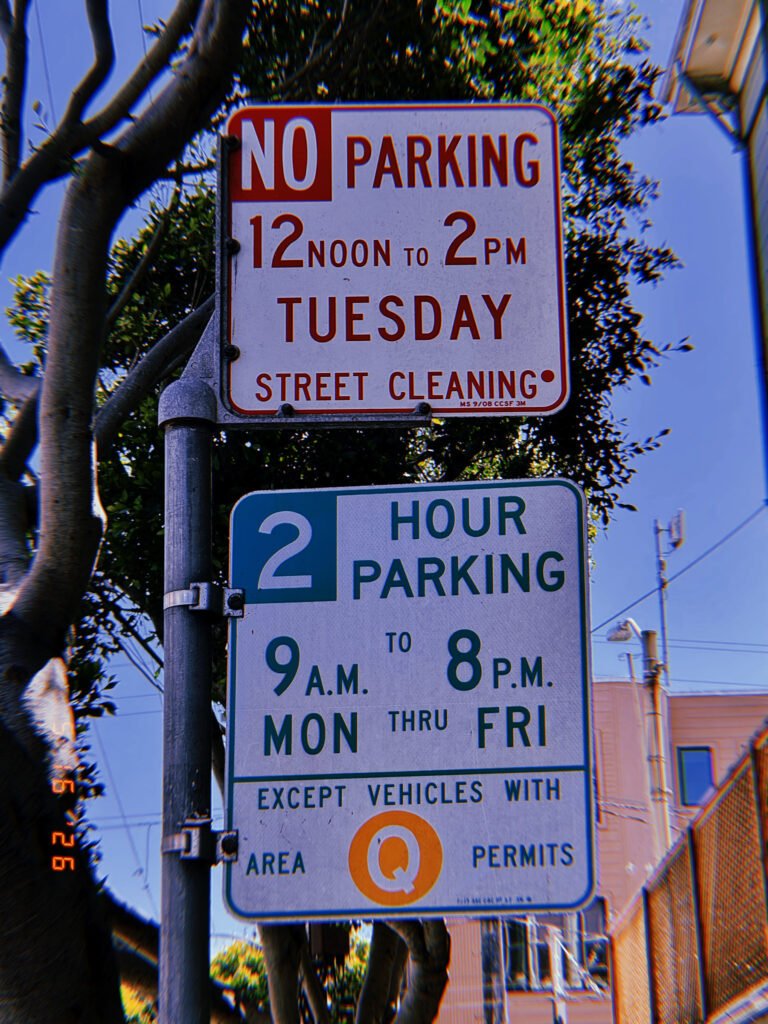 Street regulation signs mounted on a pole beside a residential street. The upper sign reserves curb access during scheduled street cleaning hours, while the lower sign limits parking duration and grants exceptions for permit holders. Together they regulate occupancy, maintenance access, and neighborhood parking allocation.