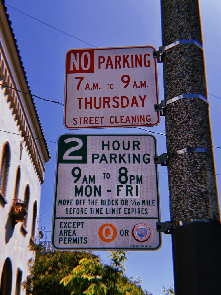 Two municipal parking regulation signs mounted on a utility pole. The upper sign restricts parking during scheduled street cleaning hours, while the lower sign establishes a two-hour parking limit with permit-holder exceptions. The assembly coordinates maintenance access and vehicle occupancy within the streetscape.