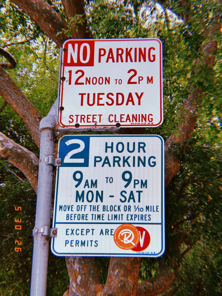 Two municipal parking regulation signs mounted on a metal pole in front of dense street trees. The upper sign prohibits parking during scheduled street cleaning hours, while the lower sign establishes a two-hour parking limit with permit-holder exemptions and a partially covered permit designation.