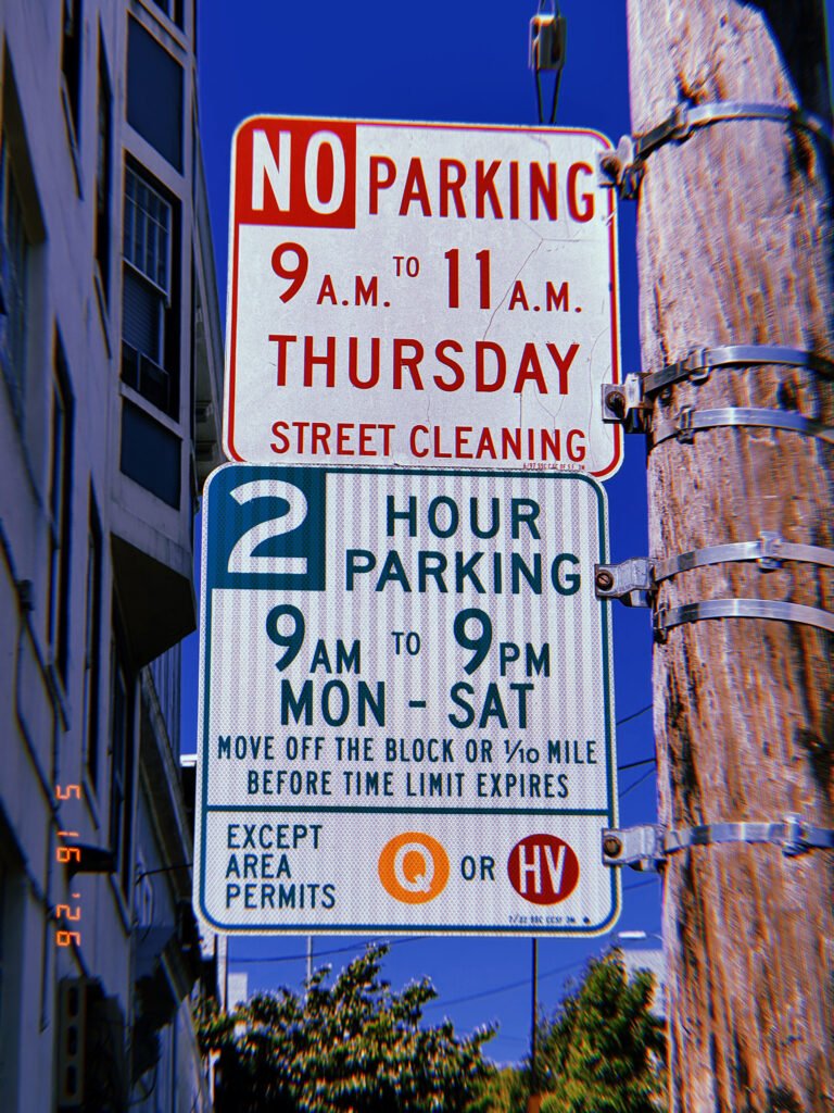 Two municipal regulation signs mounted on a utility pole. The upper sign prohibits parking during scheduled street cleaning hours, while the lower sign limits parking duration and defines permit-holder exemptions. Together they regulate maintenance access and temporary curb occupancy.