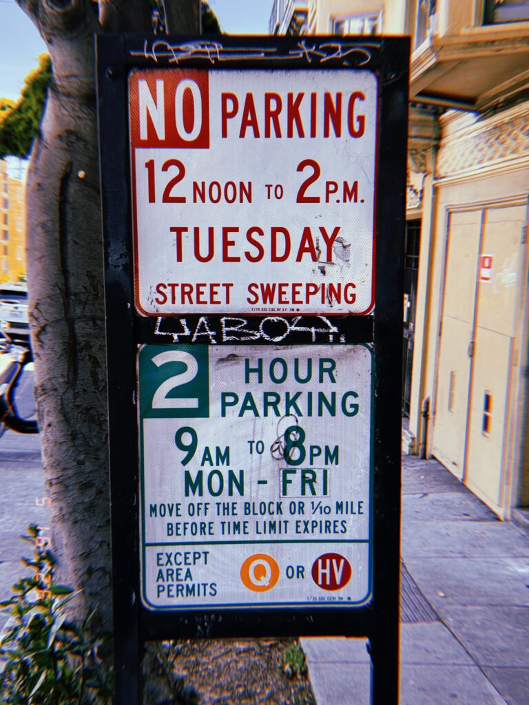 A municipal parking regulation sign displaying street sweeping restrictions, two-hour parking limits, and permit-holder exemptions. The sign is mounted within a freestanding black frame positioned along a city sidewalk beside a tree and mixed-use urban buildings.