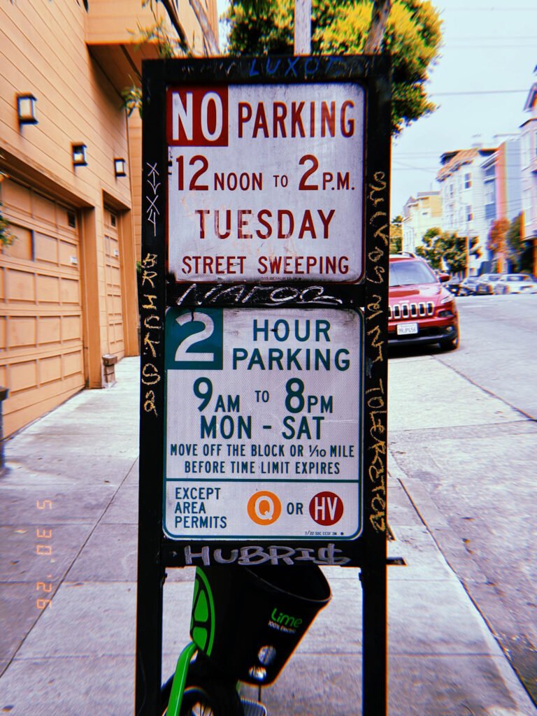 A combined municipal parking regulation sign mounted within a freestanding street fixture. The sign specifies street sweeping restrictions, time-limited parking, and permit exemptions. Graffiti markings surround the sign assembly, while a shared mobility scooter is partially visible below.