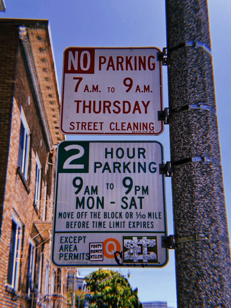 Two municipal parking regulation signs mounted on a utility pole. The upper sign restricts parking during scheduled street cleaning hours, while the lower sign establishes a two-hour parking limit with permit-holder exceptions. Stickers and markings partially cover the lower sign.