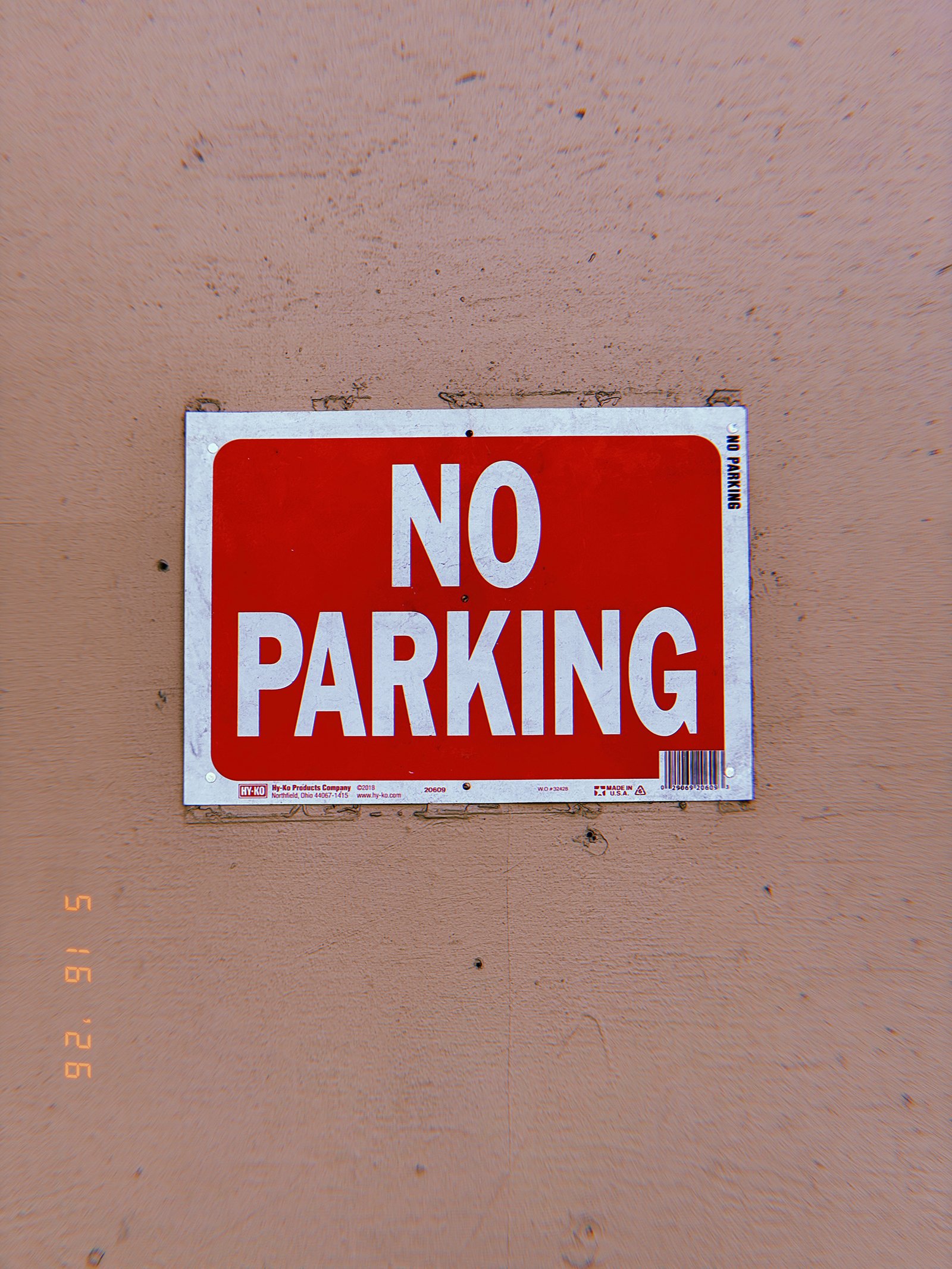 A red and white “NO PARKING” sign mounted on a plain exterior wall. Isolated from surrounding architectural features and additional instructions, the sign presents a direct restriction that marks a private access zone through a simple standardized command.