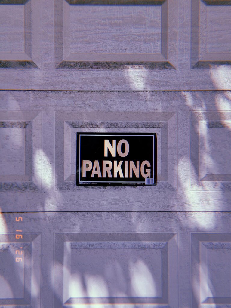 A black and white “NO PARKING” sign mounted in the recessed center panel of a residential garage door. Soft shadows from nearby trees move across the surface, while the sign remains fixed as a visual marker defining private access and restricting vehicle use of the driveway.