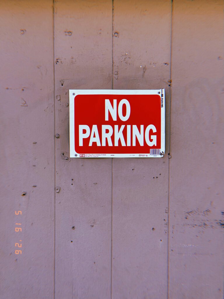 A red and white “NO PARKING” sign mounted on a plain plywood construction barrier. Centered within a large field of weathered panels and exposed fasteners, the sign functions as a direct access restriction with no additional explanation or qualification.