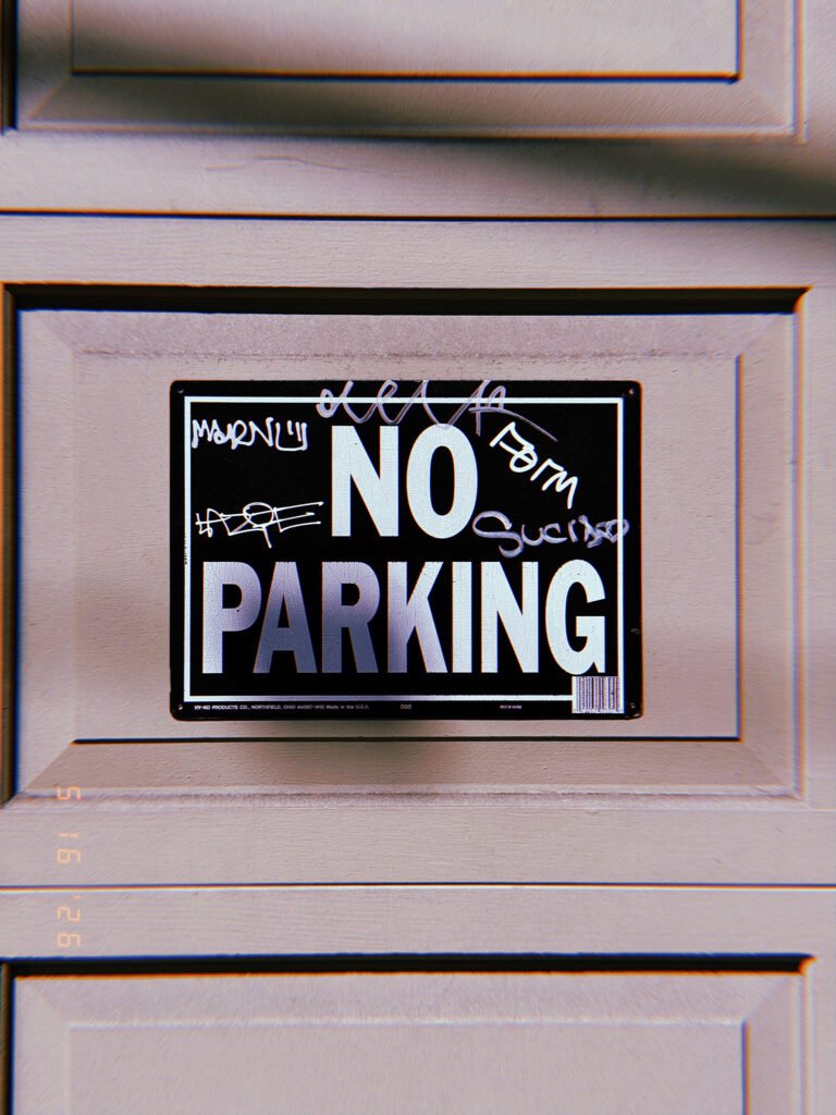 A black and white “NO PARKING” sign mounted within a recessed panel of a residential garage door. Layers of handwritten graffiti partially cover the sign’s surface, altering but not obscuring the original instruction that reserves the driveway area for property access.
