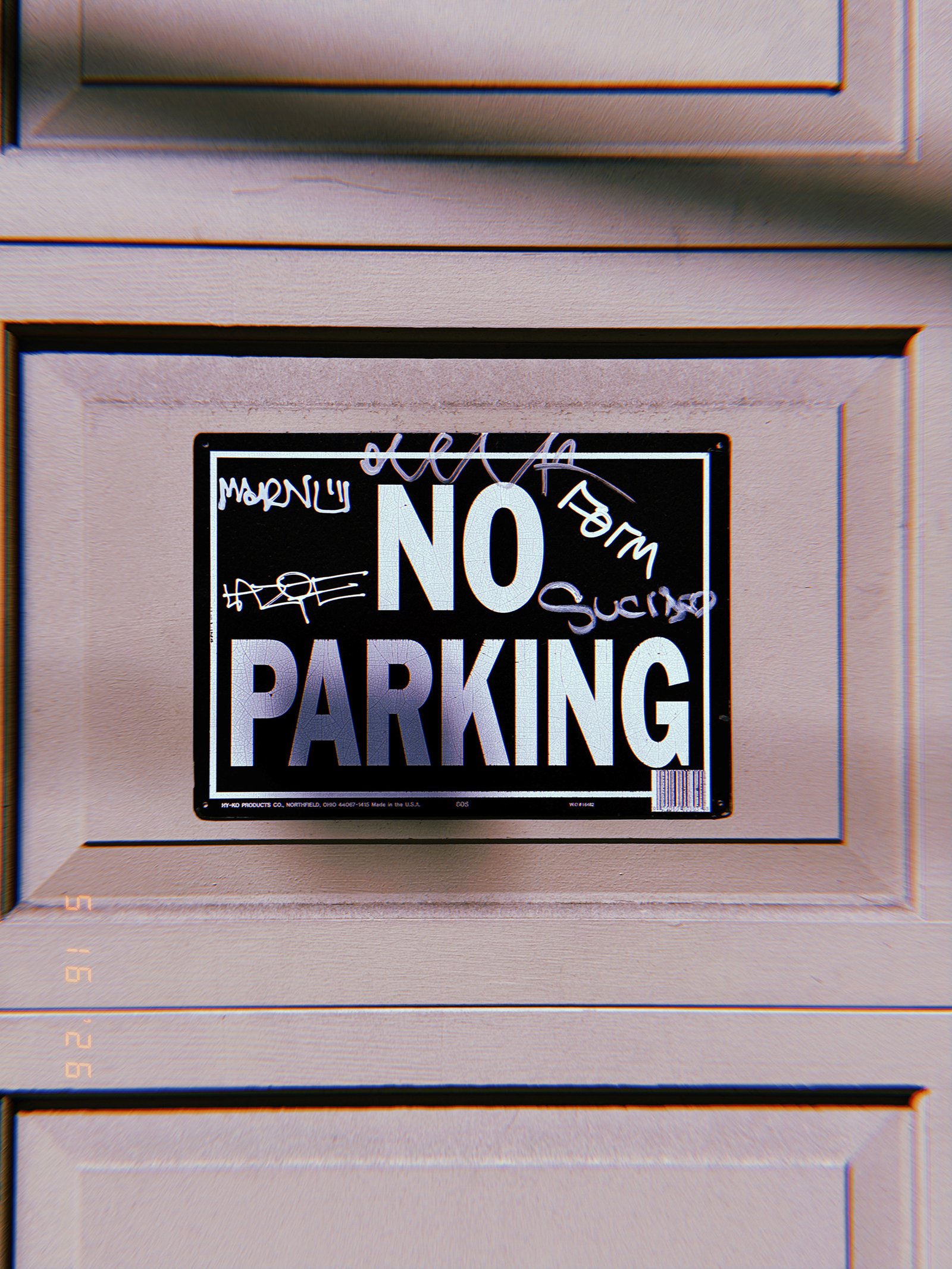 A black and white “NO PARKING” sign mounted within a recessed panel of a residential garage door. Layers of handwritten graffiti partially cover the sign’s surface, altering but not obscuring the original instruction that reserves the driveway area for property access.