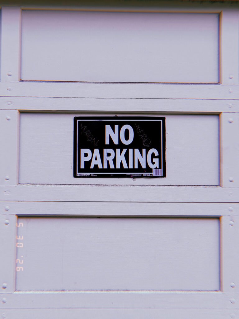 A black and white “NO PARKING” sign mounted at the center of a residential garage door. The sign establishes a private access restriction, reserving the driveway opening and preventing vehicles from obstructing entry to the property.