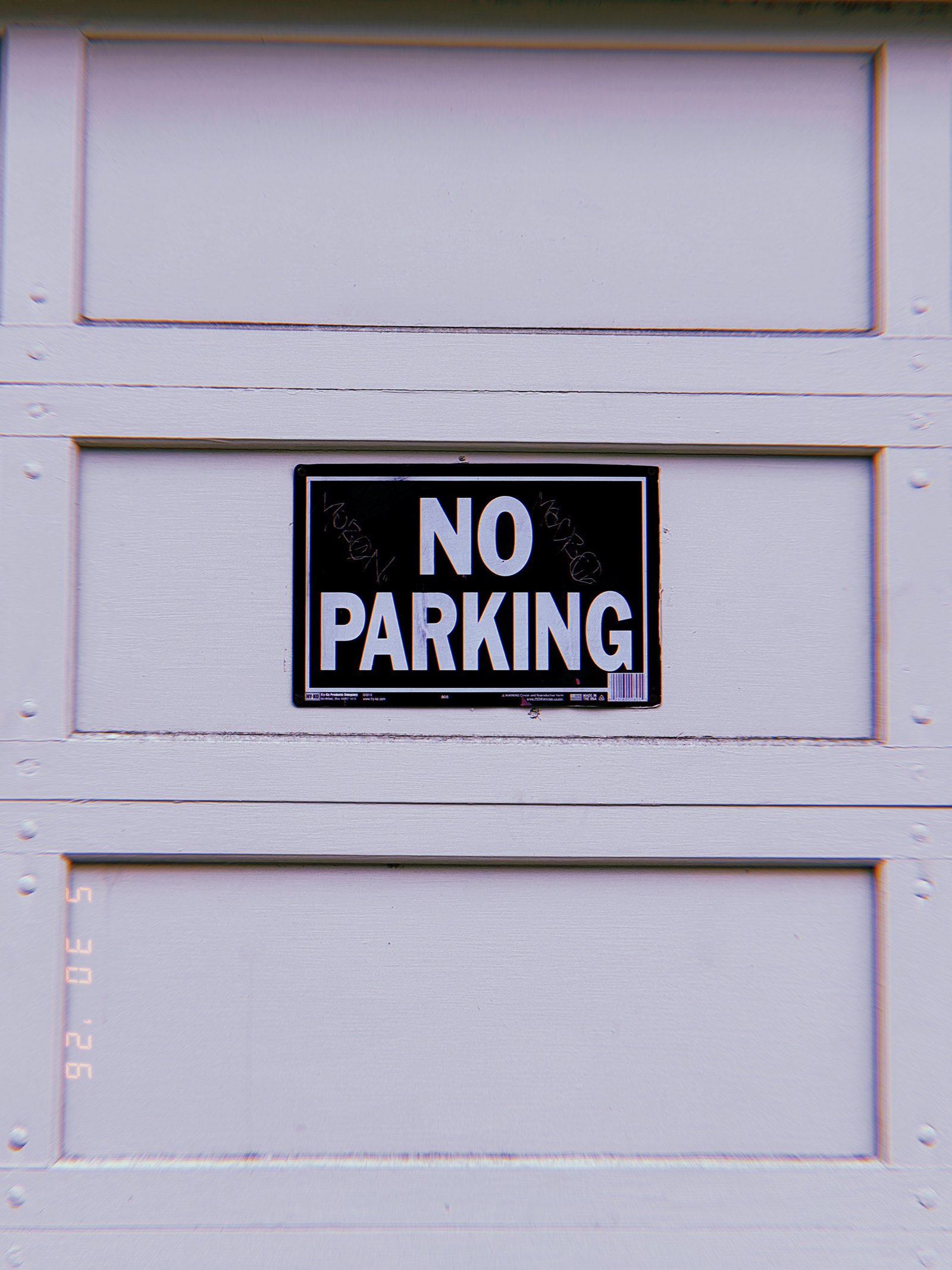 A black and white “NO PARKING” sign mounted at the center of a residential garage door. The sign establishes a private access restriction, reserving the driveway opening and preventing vehicles from obstructing entry to the property.