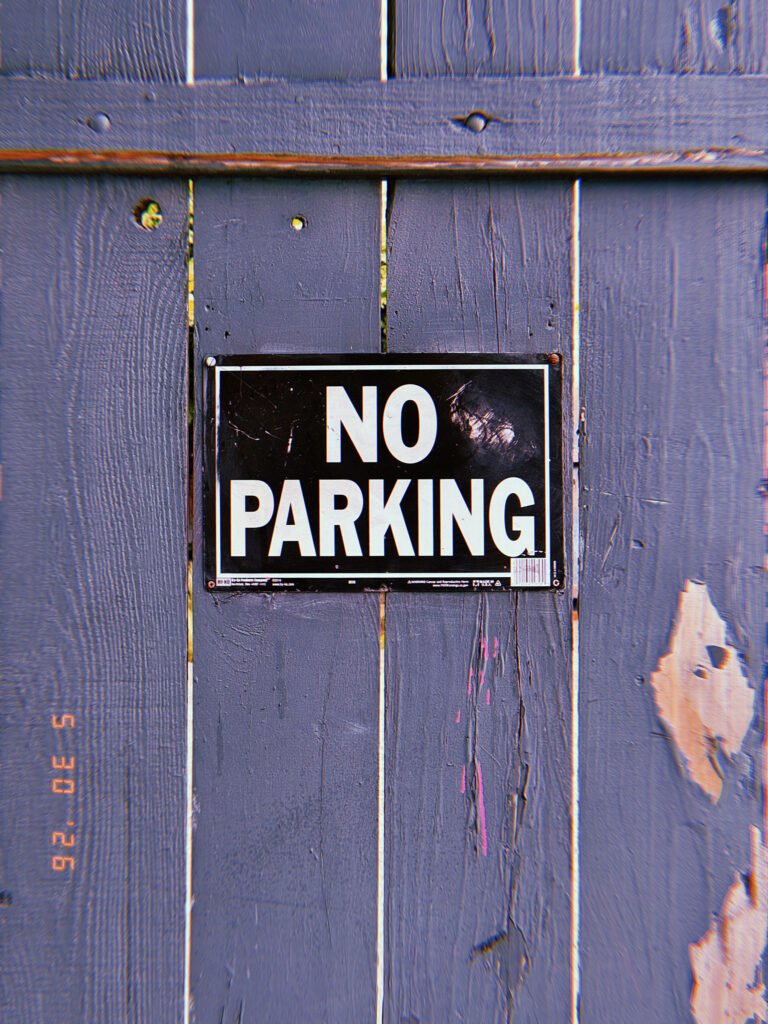 A black and white “NO PARKING” sign mounted on a weathered wooden fence. The sign establishes a private access restriction, preventing vehicle occupancy along the boundary and preserving access to the property beyond.