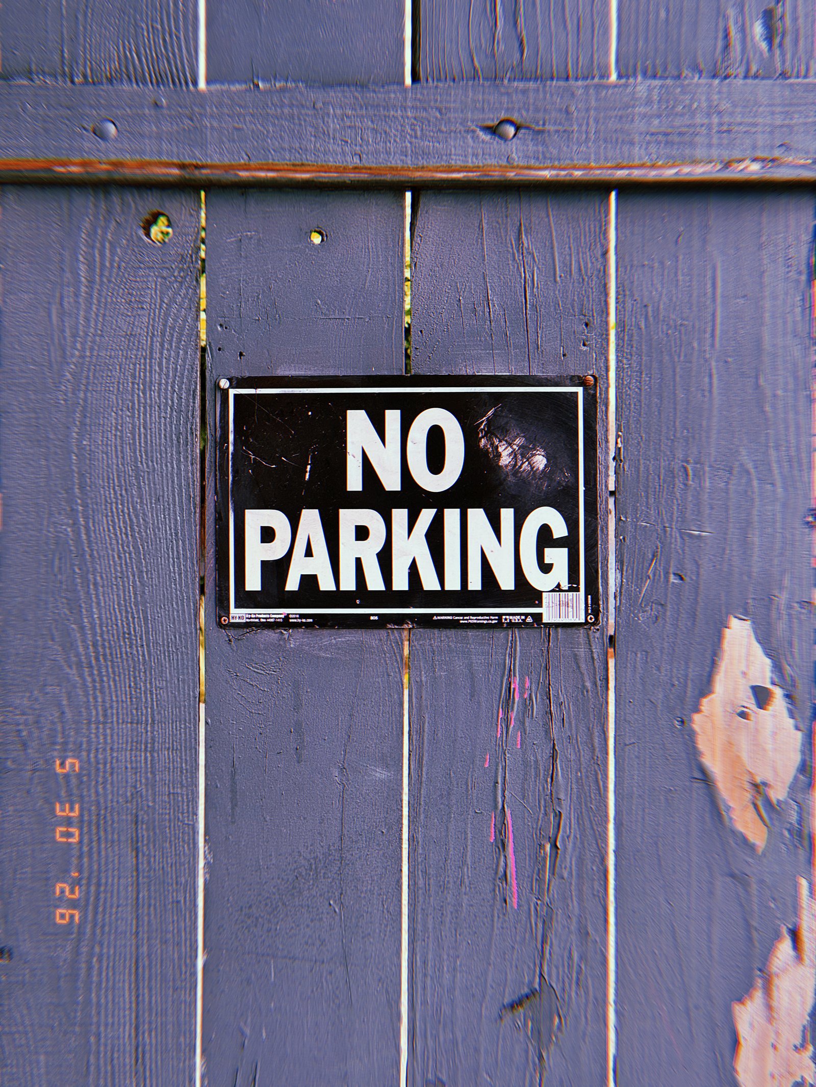 A black and white “NO PARKING” sign mounted on a weathered wooden fence. The sign establishes a private access restriction, preventing vehicle occupancy along the boundary and preserving access to the property beyond.