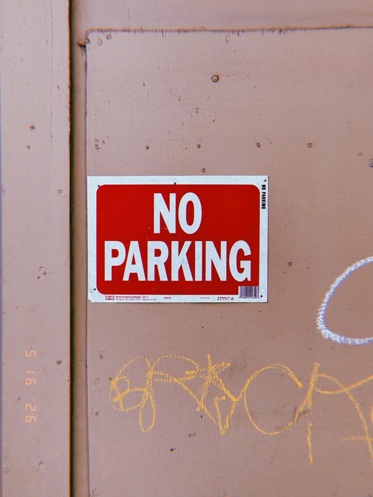 A red and white “NO PARKING” sign mounted on a flat wall surface. The sign establishes a localized parking restriction at the edge of private property, reserving adjacent space for access, circulation, or operational use.