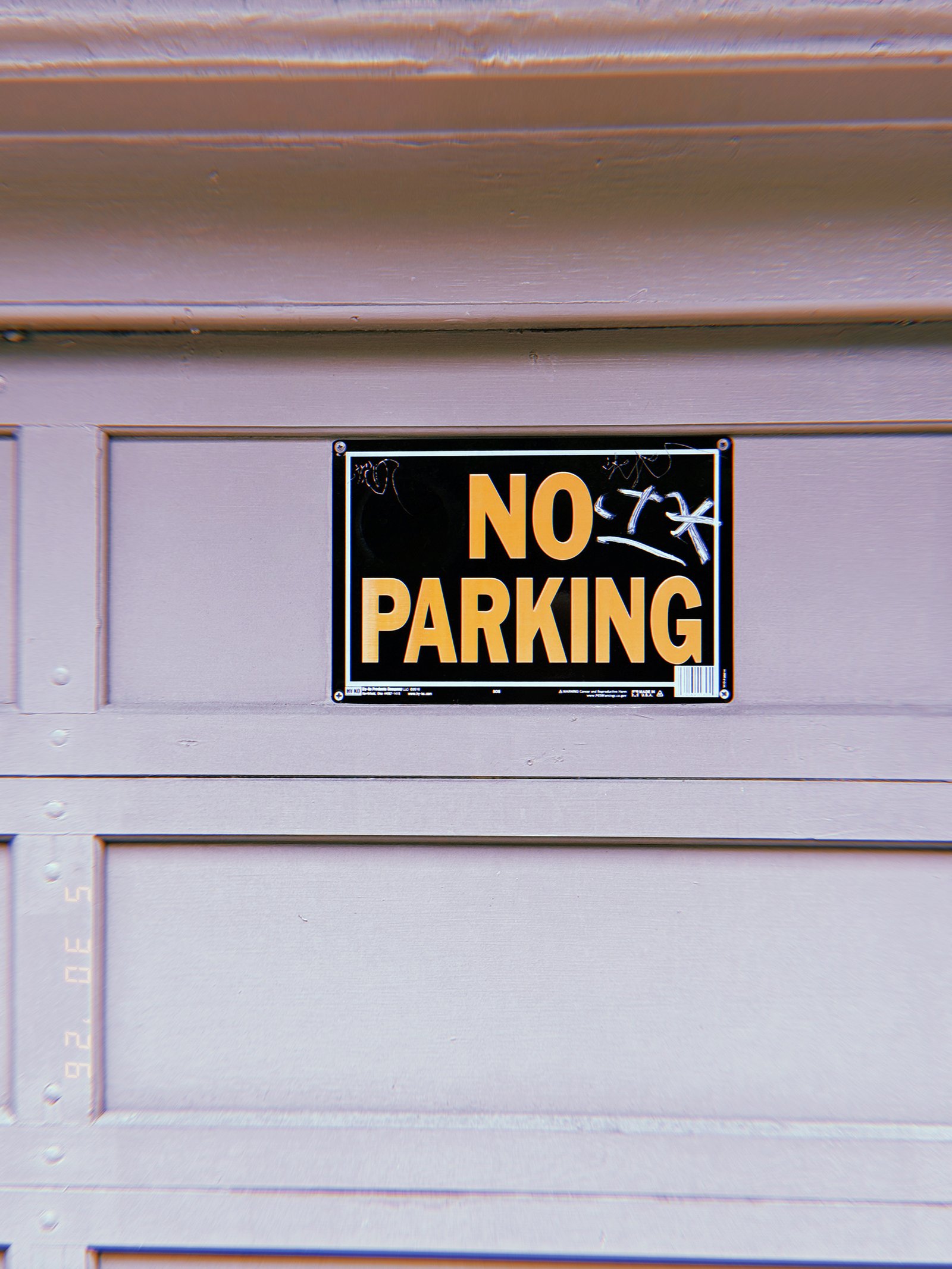 A small “NO PARKING” sign mounted on a residential garage door. The sign establishes a localized access restriction, reserving the driveway entrance and preventing vehicles from obstructing entry to private property.