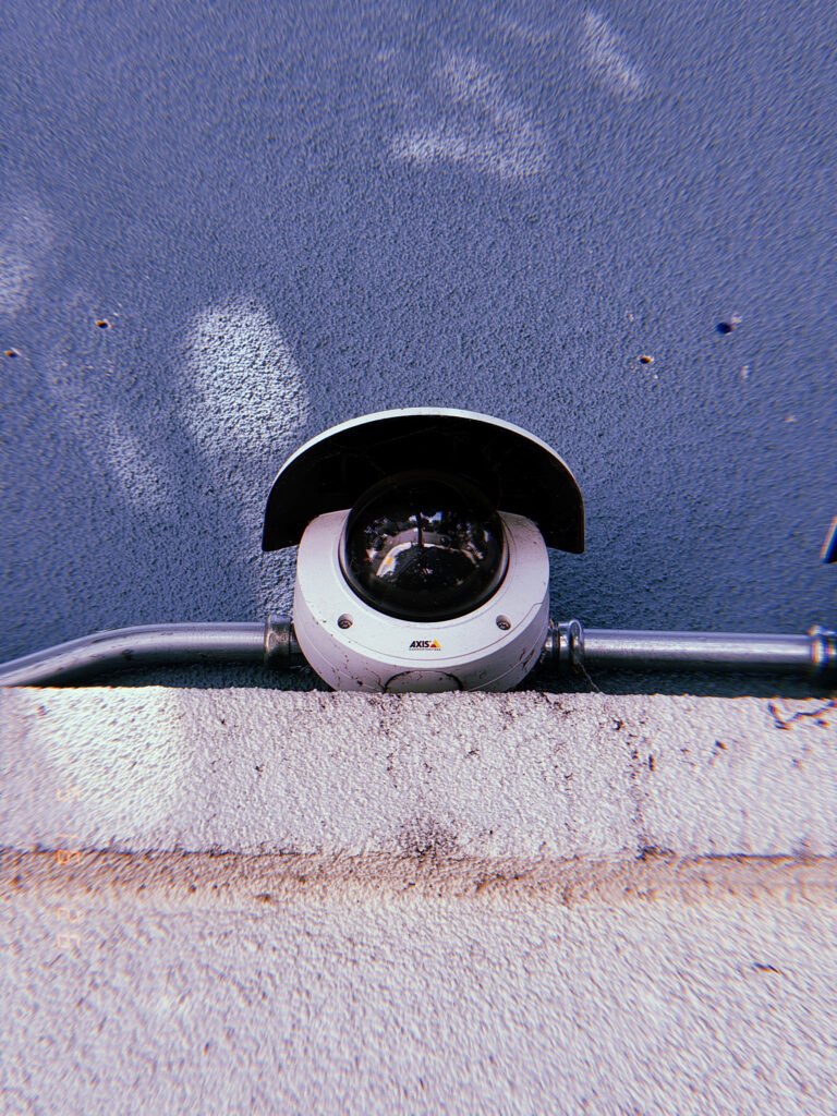 A dome surveillance camera mounted behind a protective hood and connected to exposed metal conduit. Installed along a wall junction, the device forms part of the built observation infrastructure embedded within the urban environment.