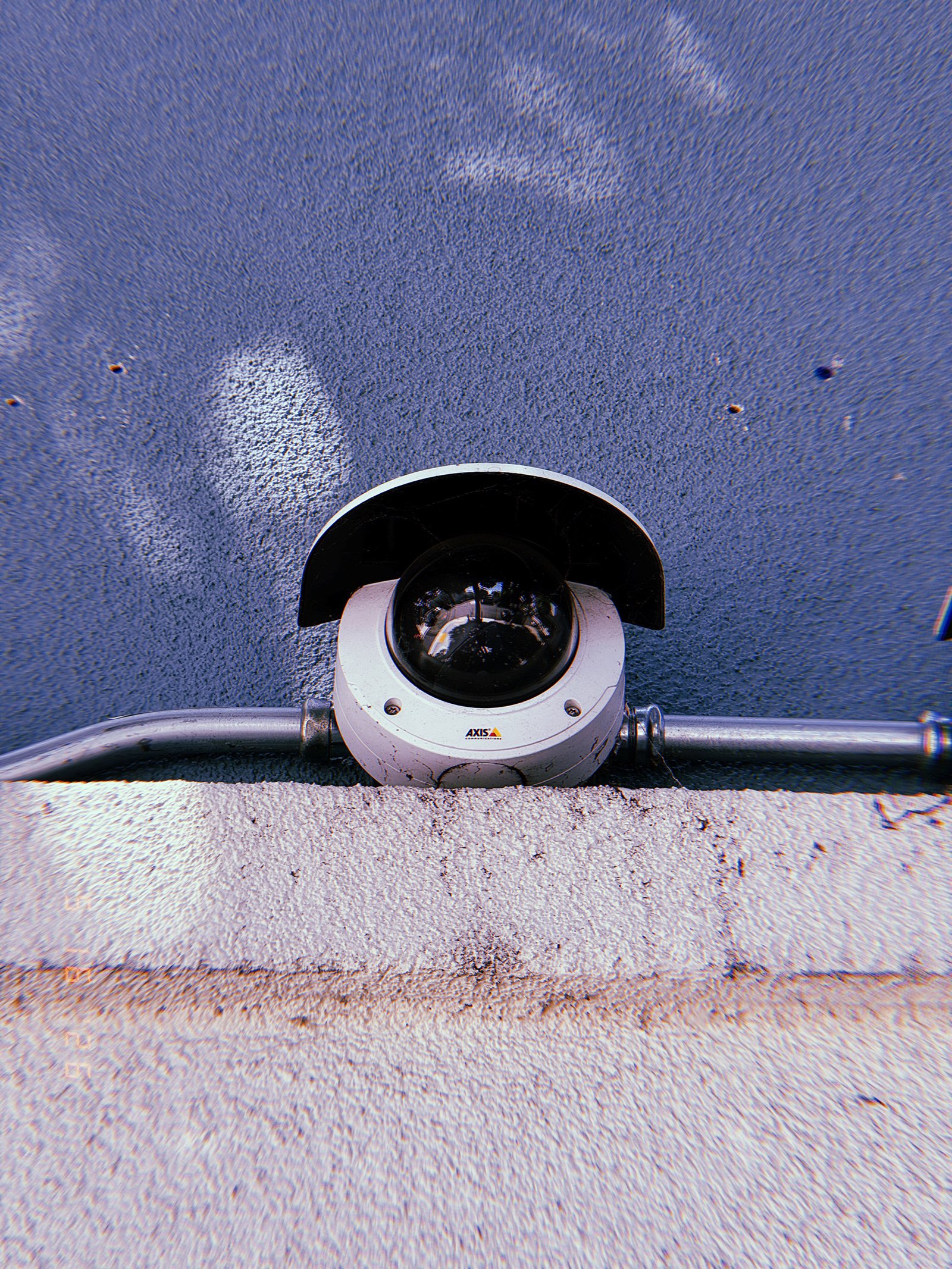 A dome surveillance camera mounted behind a protective hood and connected to exposed metal conduit. Installed along a wall junction, the device forms part of the built observation infrastructure embedded within the urban environment.