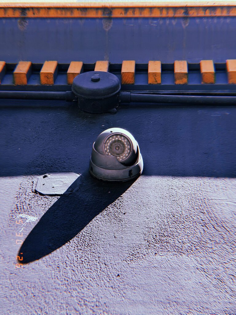 A weathered surveillance camera mounted on a dark rooftop surface. Strong sunlight casts a long shadow across the structure, while cables and utility components above connect the device to the surrounding observation network.
