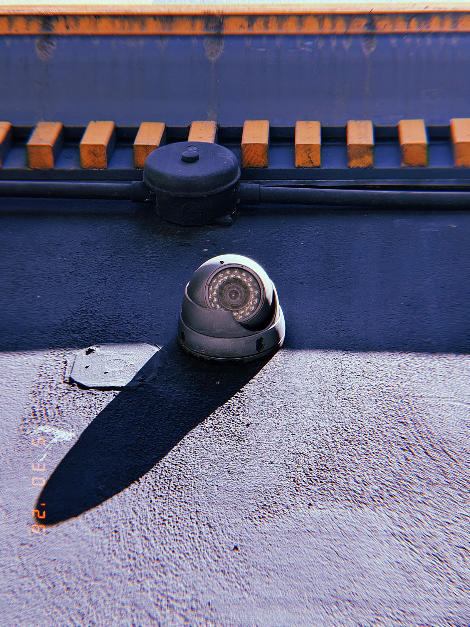 A weathered surveillance camera mounted on a dark rooftop surface. Strong sunlight casts a long shadow across the structure, while cables and utility components above connect the device to the surrounding observation network.