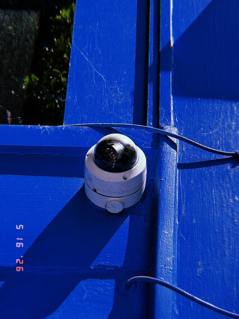 A dome surveillance camera mounted at the corner of a bright blue painted structure. Exposed cables run across the surface while strong sunlight casts angular shadows that emphasize the device’s integration into the built environment.