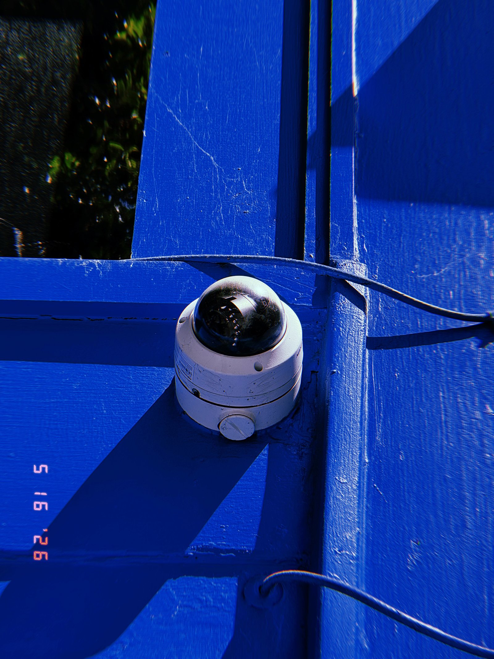 A dome surveillance camera mounted at the corner of a bright blue painted structure. Exposed cables run across the surface while strong sunlight casts angular shadows that emphasize the device’s integration into the built environment.