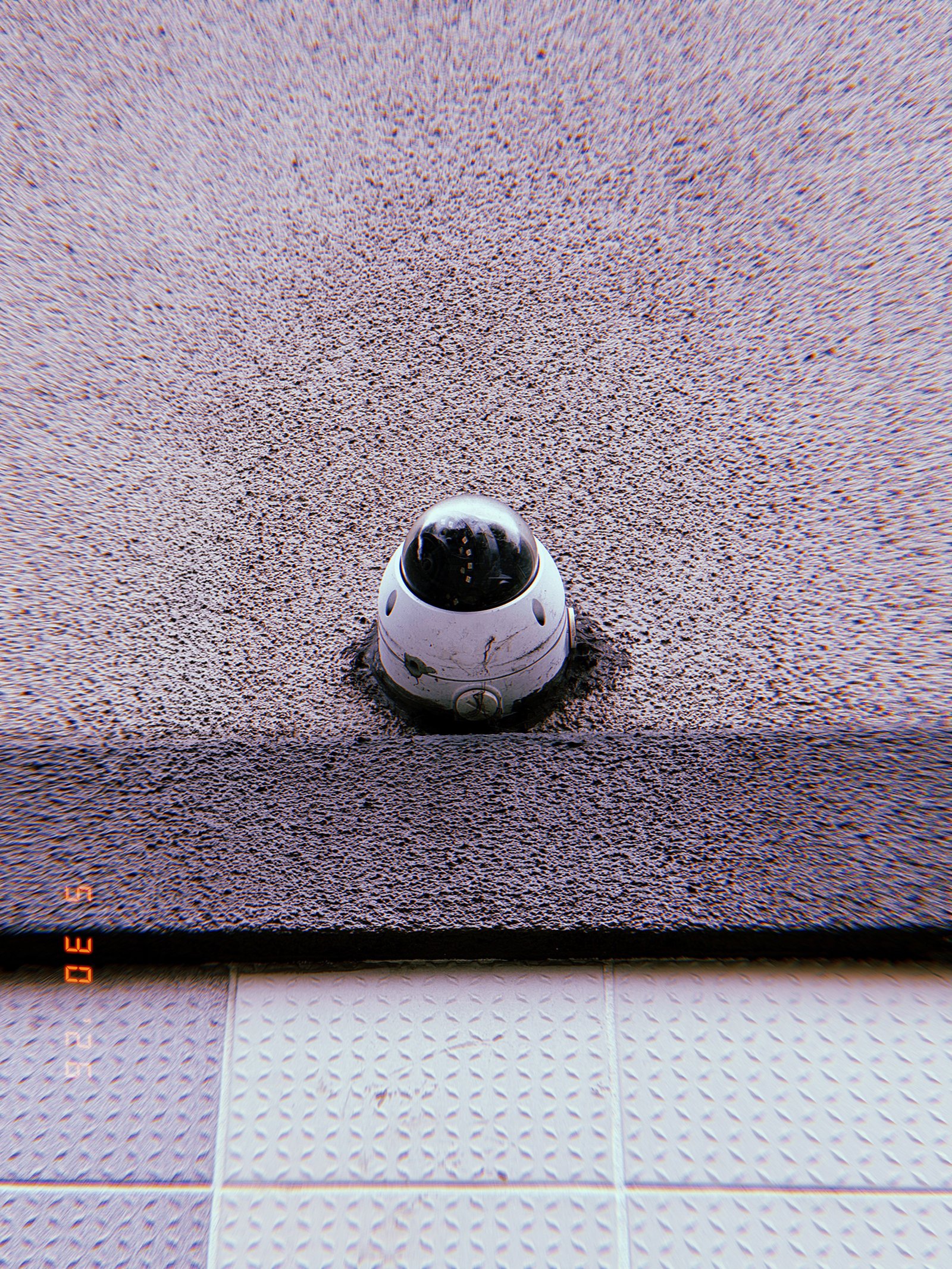 A small dome surveillance camera embedded in a textured exterior wall above a tiled surface. Positioned at the boundary between two materials, the device forms a compact observation point within the building facade.