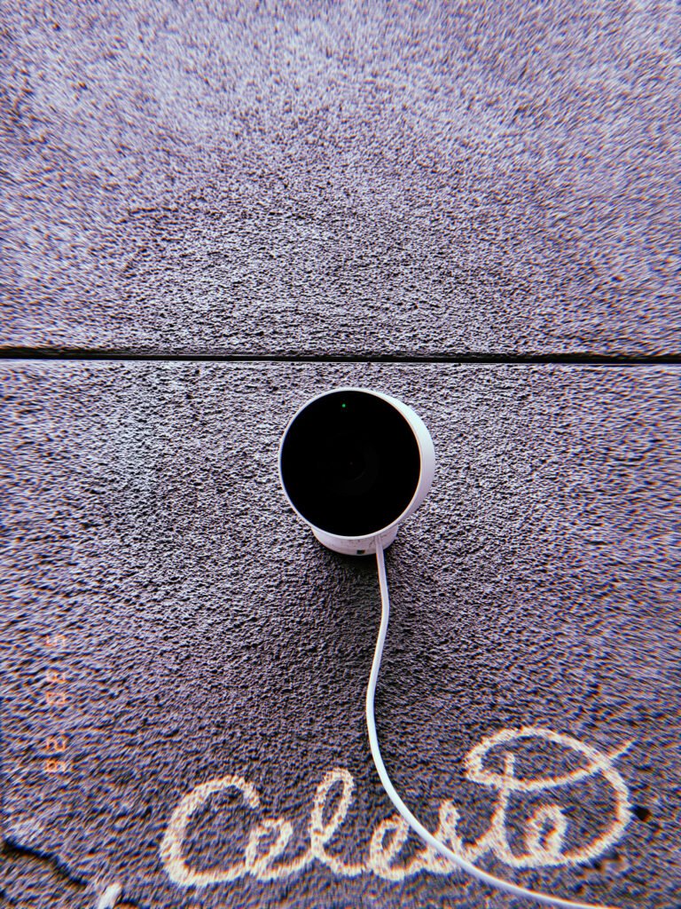 A small surveillance camera mounted on a textured exterior wall. A visible cable runs from the device across the surface, while graffiti markings below contrast with the clean geometric form of the monitoring equipment.
