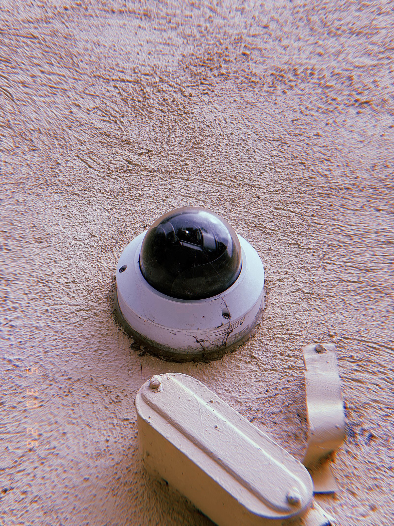 A weathered dome surveillance camera mounted on a textured exterior wall. The protective housing shows signs of age and wear, while utility hardware below connects the device to the surrounding monitoring infrastructure.