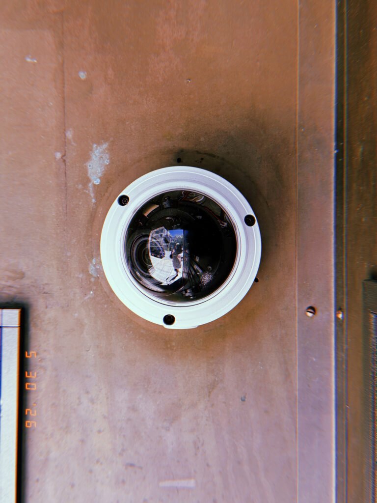 A dome surveillance camera mounted on a building facade. The transparent housing reveals internal components and lens elements, while reflections of the surrounding street appear across the curved surface of the protective dome.