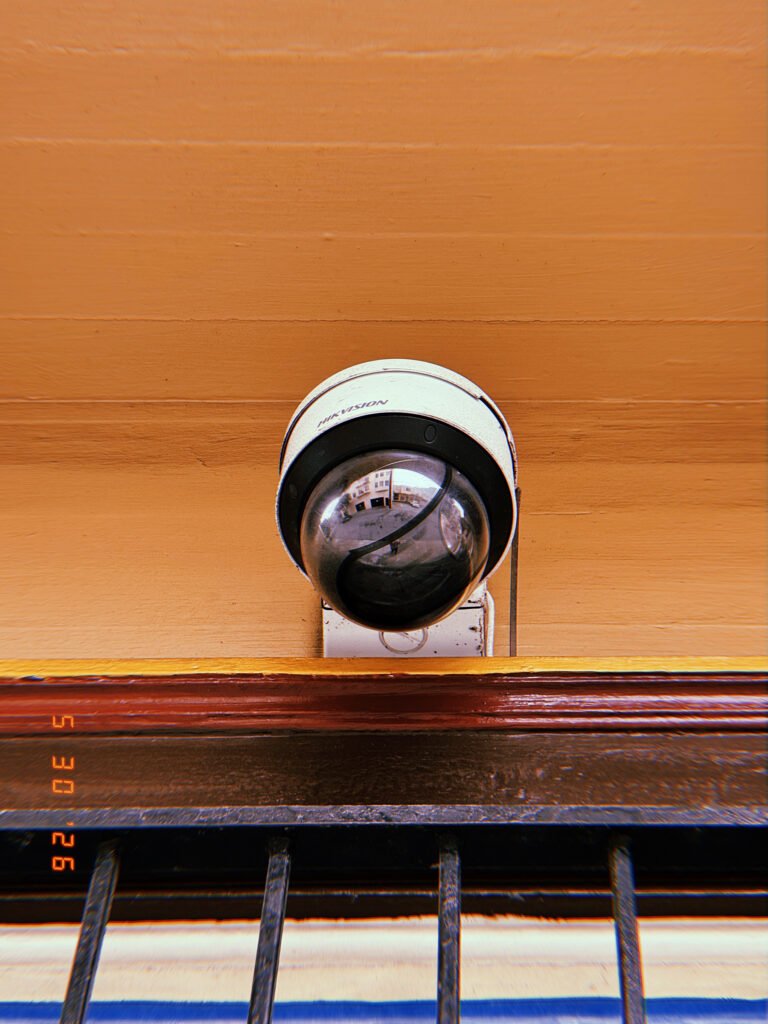 A dome surveillance camera mounted above a barred window on a building facade. The reflective housing captures fragments of the surrounding street while the device oversees the entrance area from an elevated position.