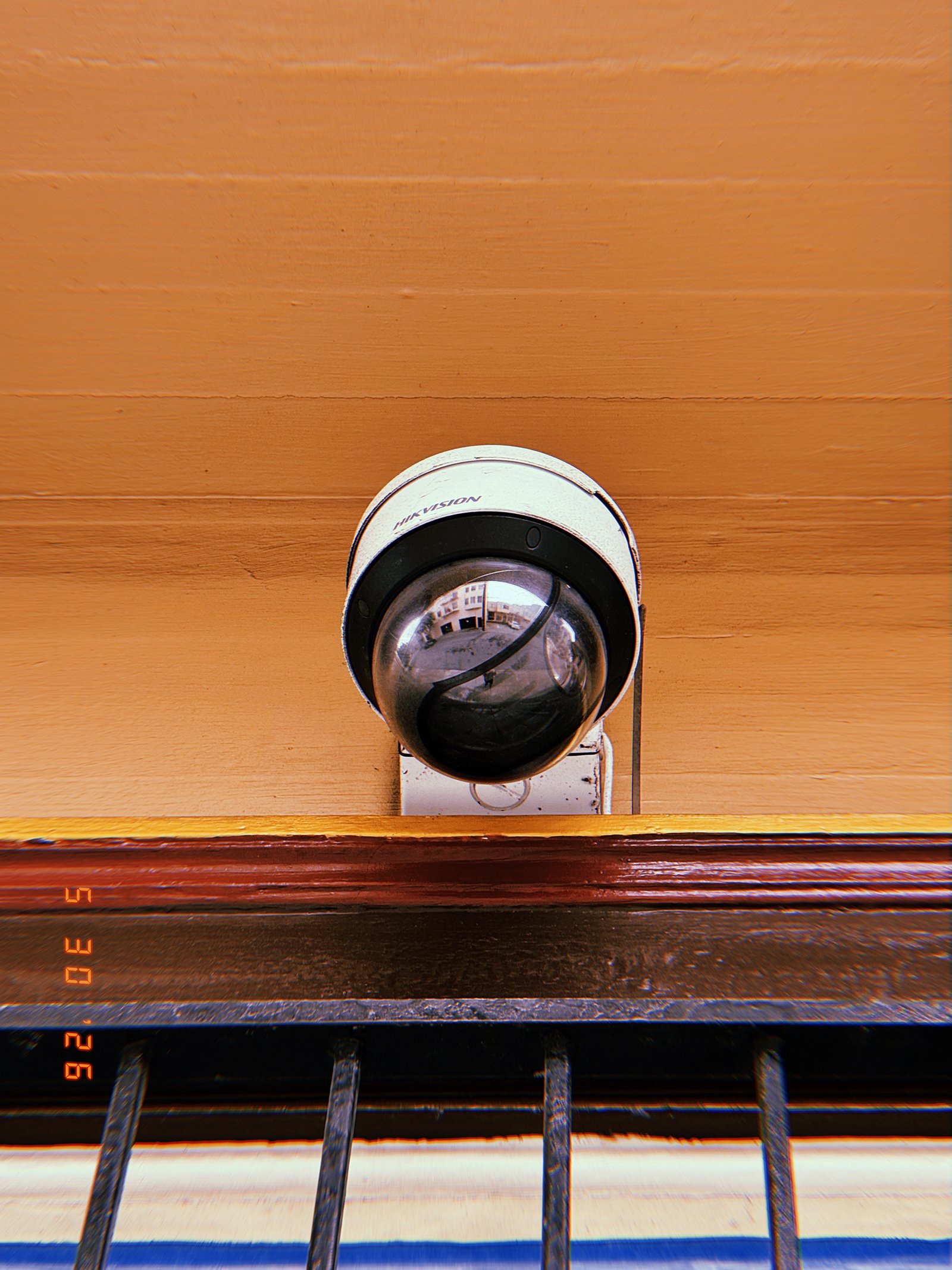 A dome surveillance camera mounted above a barred window on a building facade. The reflective housing captures fragments of the surrounding street while the device oversees the entrance area from an elevated position.