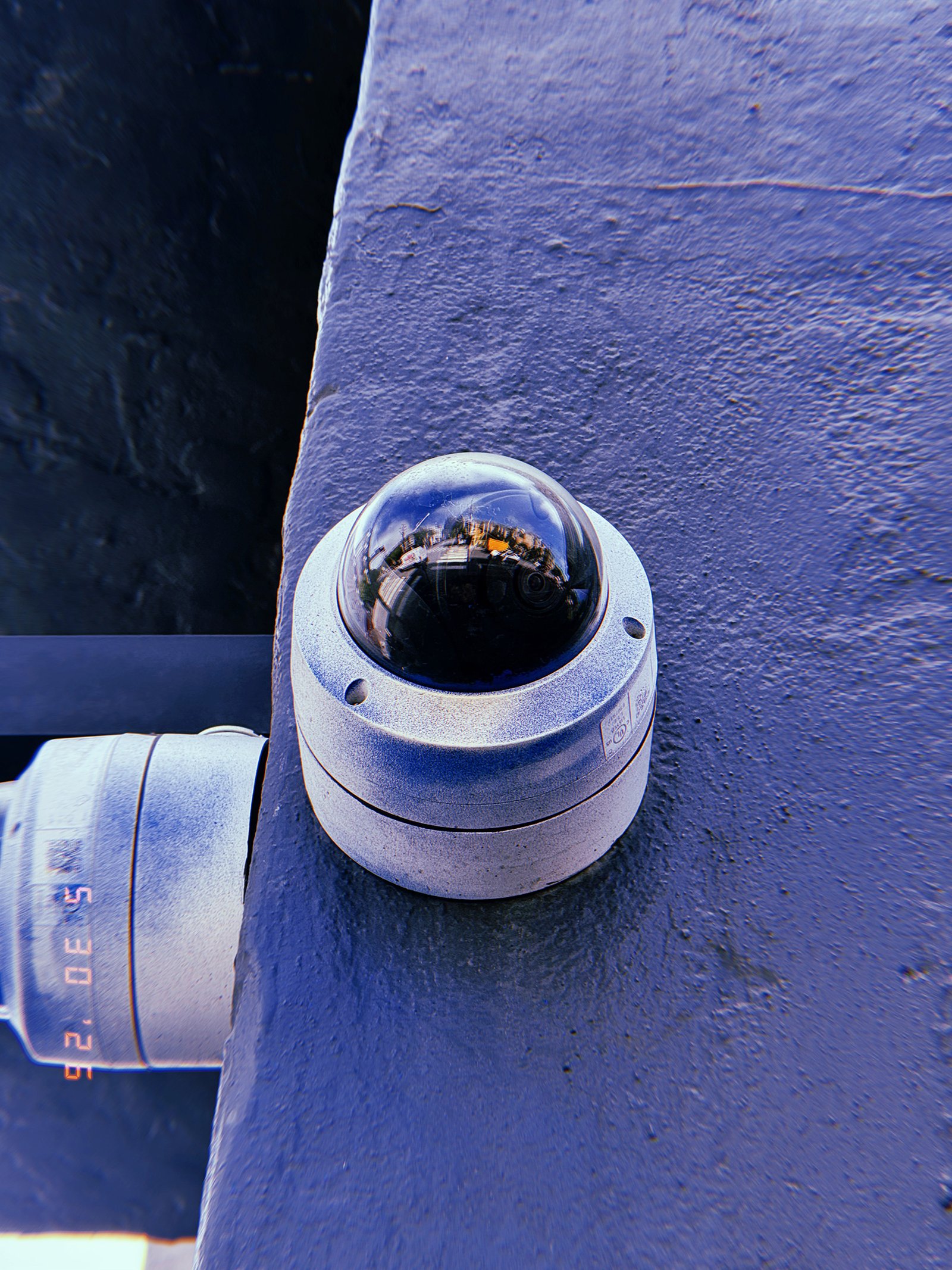 A dome-style surveillance camera mounted on the corner of a building. The reflective housing mirrors the surrounding street environment while the device occupies a strategic position overlooking adjacent public space.