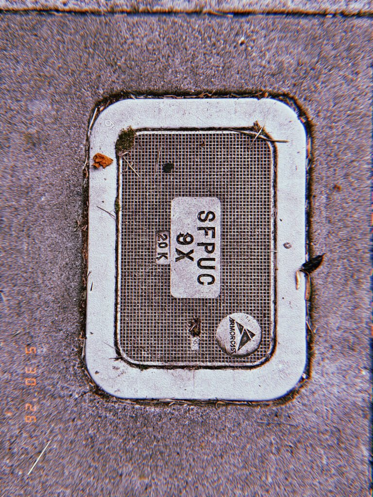 A rectangular communications access cover embedded in a concrete sidewalk. The textured utility panel contains identification markings and provides maintenance access to underground communication infrastructure.