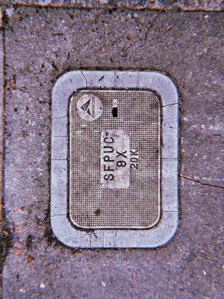 A rectangular communications access cover embedded in a concrete sidewalk. The textured utility panel contains service markings and provides maintenance access to underground communication infrastructure.
