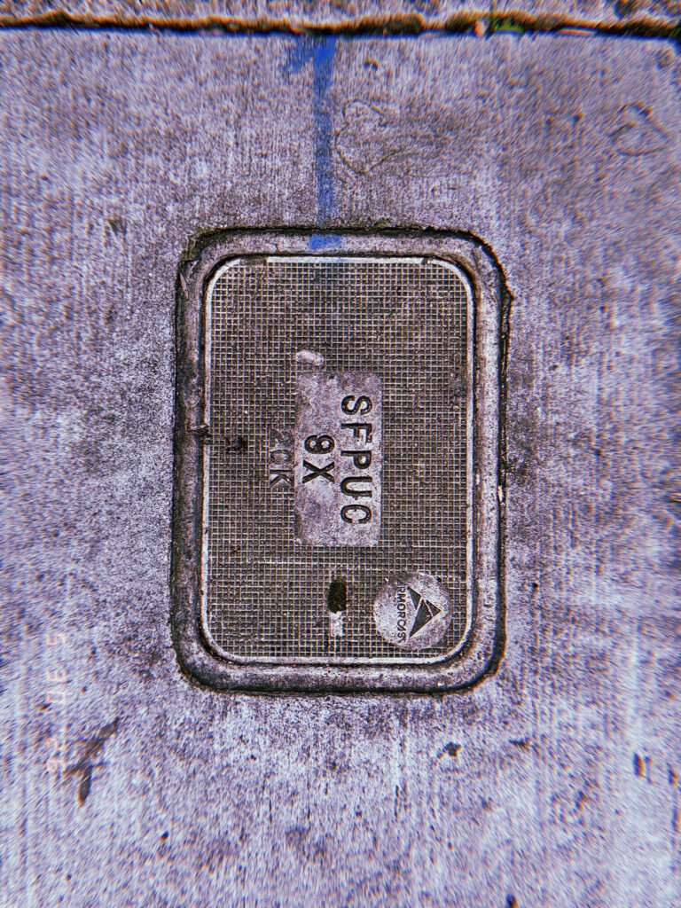 A rectangular communications access cover embedded within a concrete sidewalk. The textured utility panel contains identification markings and provides access to underground communication infrastructure.