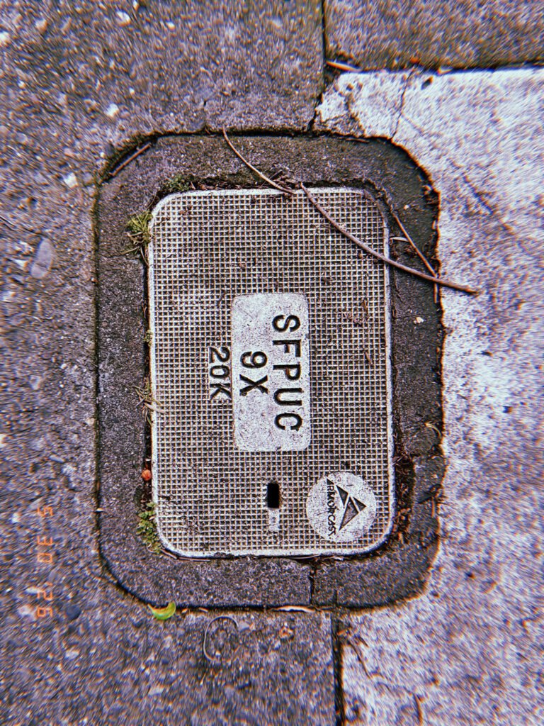 A rectangular communications access cover set into the edge of a sidewalk and asphalt surface. Utility markings and identification labels indicate access to underground network infrastructure.