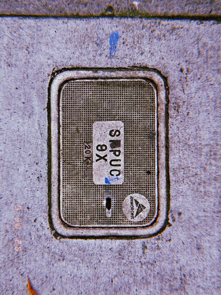 A rectangular utility access cover embedded in a concrete sidewalk. The textured lid carries service markings and identification labels, forming a maintenance access point to underground communications infrastructure.