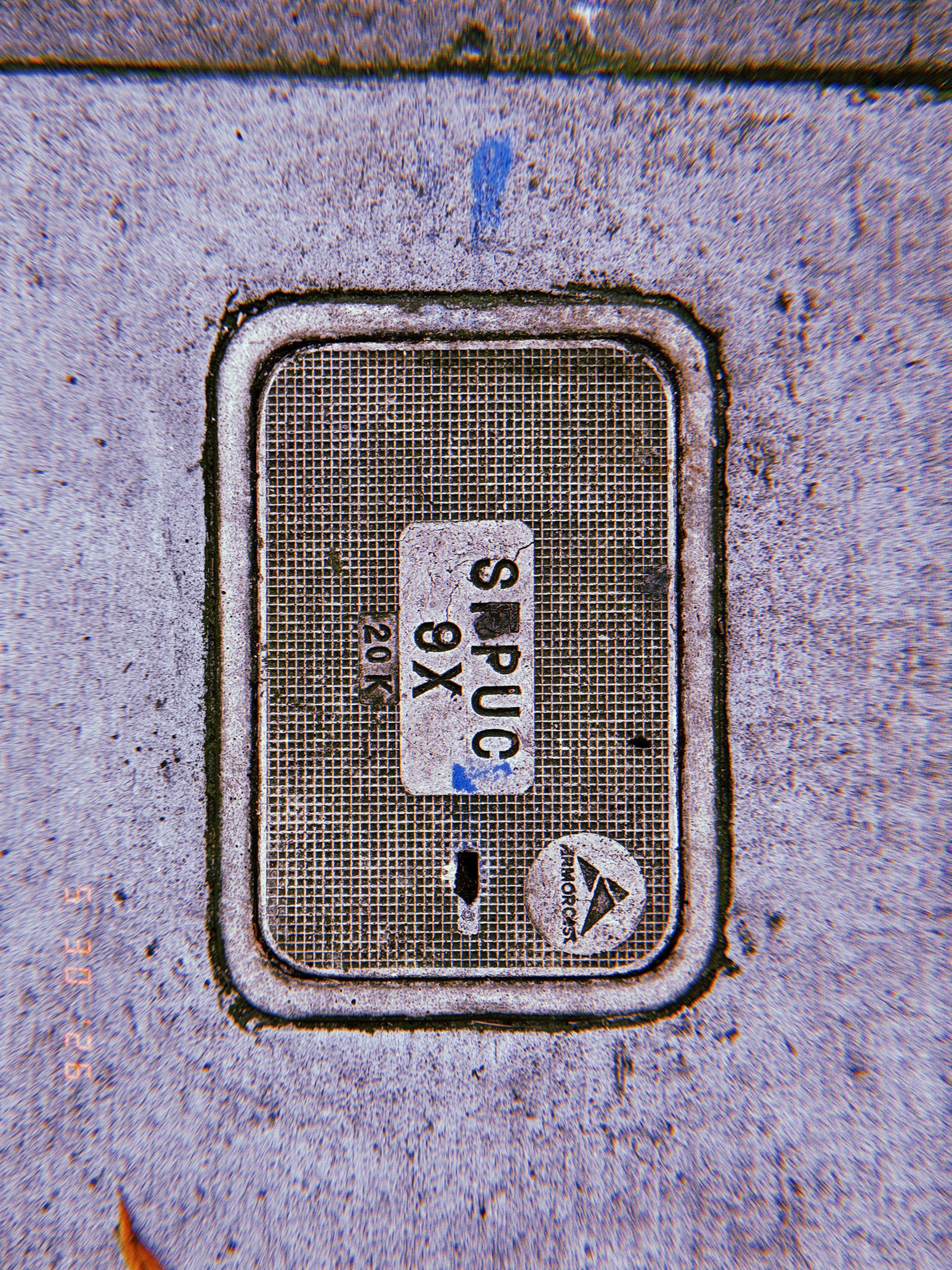 A rectangular utility access cover embedded in a concrete sidewalk. The textured lid carries service markings and identification labels, forming a maintenance access point to underground communications infrastructure.