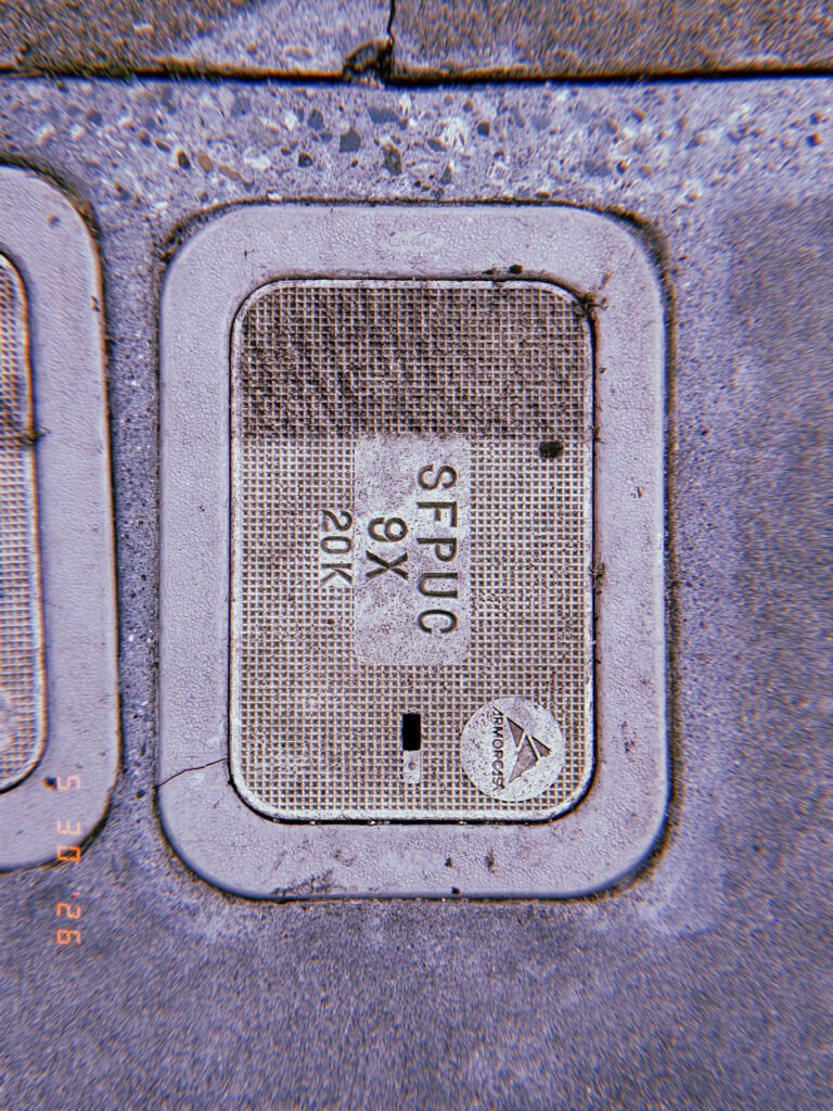A rectangular utility access cover embedded in a concrete sidewalk. The textured lid is marked with service identifiers and surrounded by weathered pavement, providing access to underground communications infrastructure.