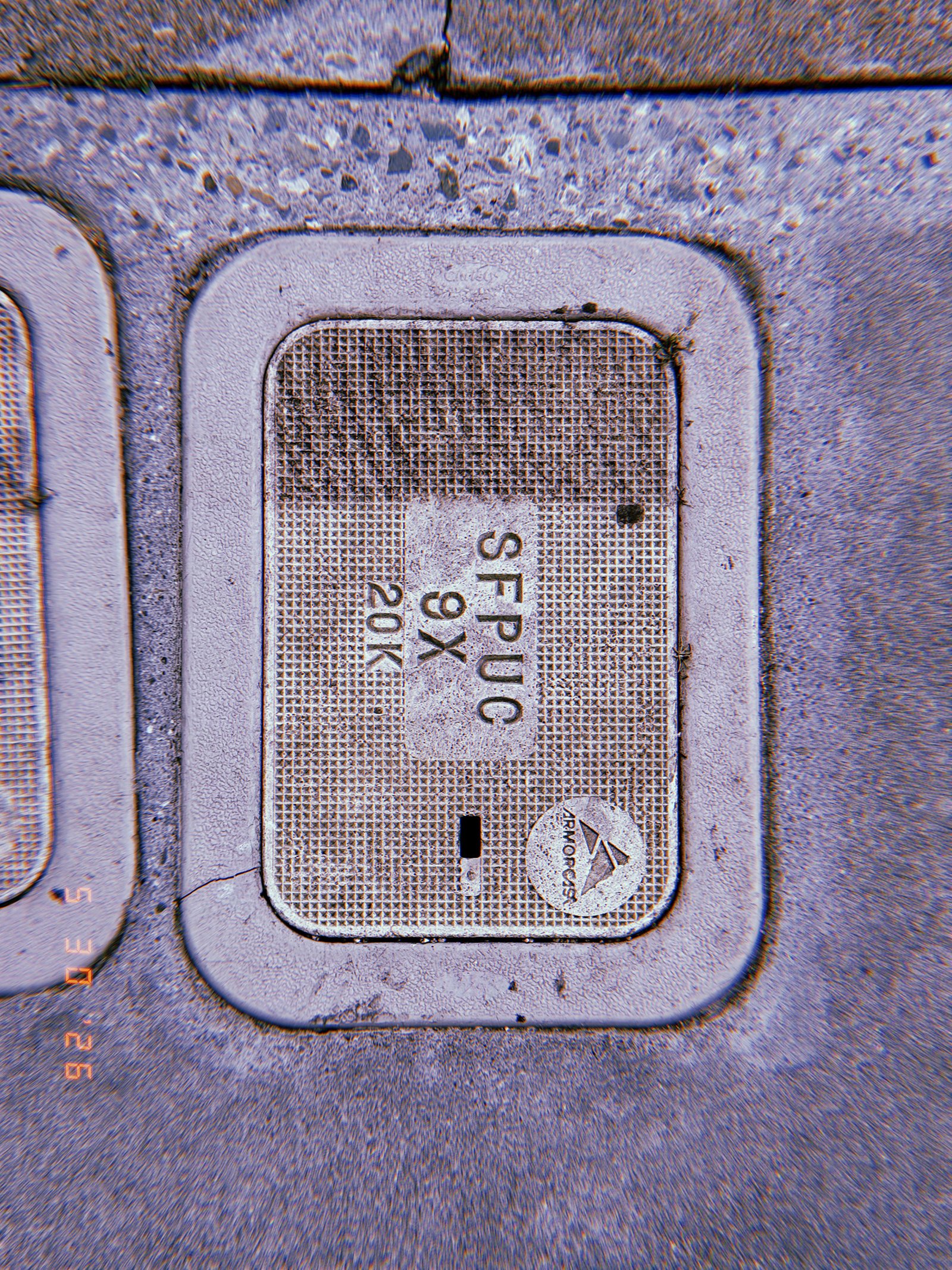 A rectangular utility access cover embedded in a concrete sidewalk. The textured lid is marked with service identifiers and surrounded by weathered pavement, providing access to underground communications infrastructure.