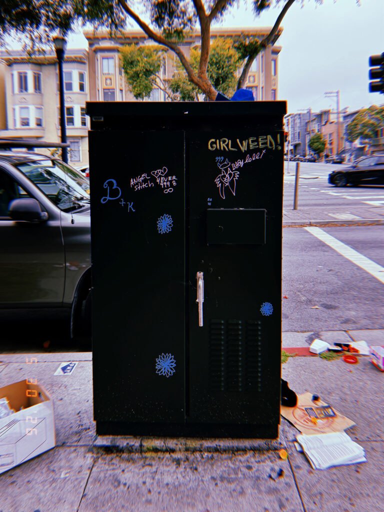 A black utility cabinet on a San Francisco sidewalk covered with graffiti, stickers, and handwritten markings, enclosing electrical or communications infrastructure.