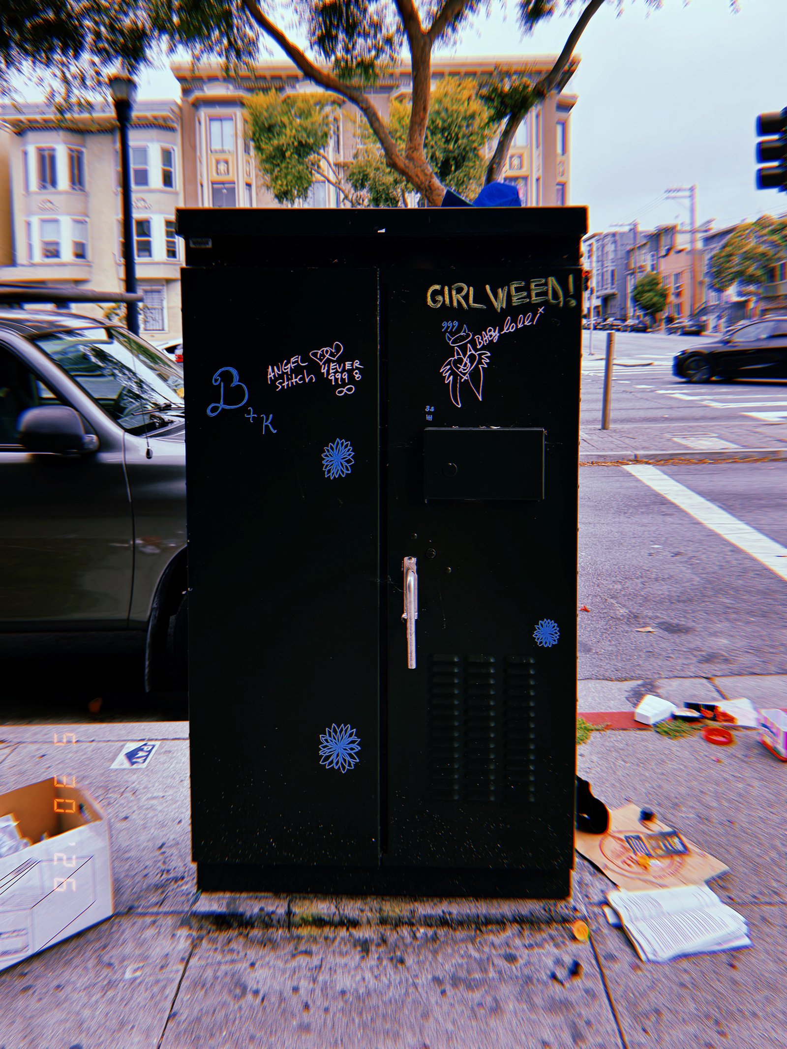 A black utility cabinet on a San Francisco sidewalk covered with graffiti, stickers, and handwritten markings, enclosing electrical or communications infrastructure.