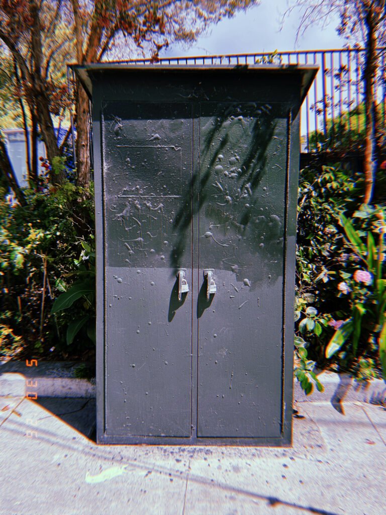 A tall gray utility cabinet with double locking doors installed along a San Francisco sidewalk, protecting electrical or communications infrastructure behind a secure metal enclosure.